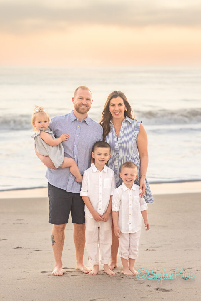 A family of five stands on a beach at sunset. The father holds a toddler. Two young boys stand in front, all wearing light-colored clothes. The mother stands beside them, smiling. Waves and a pastel sky form the backdrop. Vero Beach Photoshoot