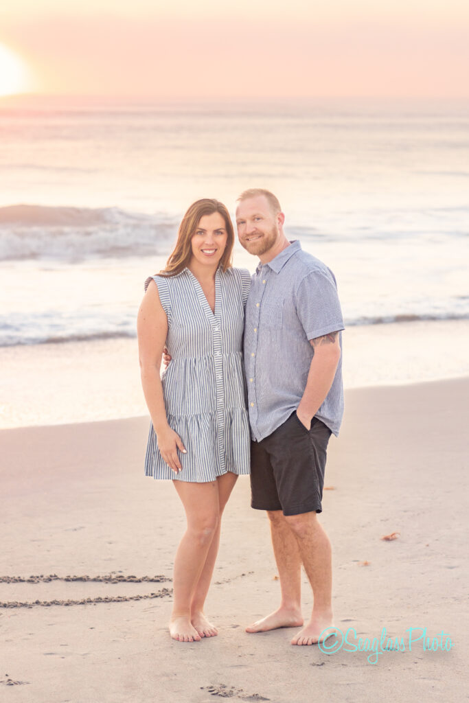 A couple stands barefoot on a beach during sunset, with the ocean waves in the background. The woman wears a striped dress, and the man wears a short-sleeve button-up shirt and shorts. They are smiling and embracing each other. Vero Beach Photoshoot