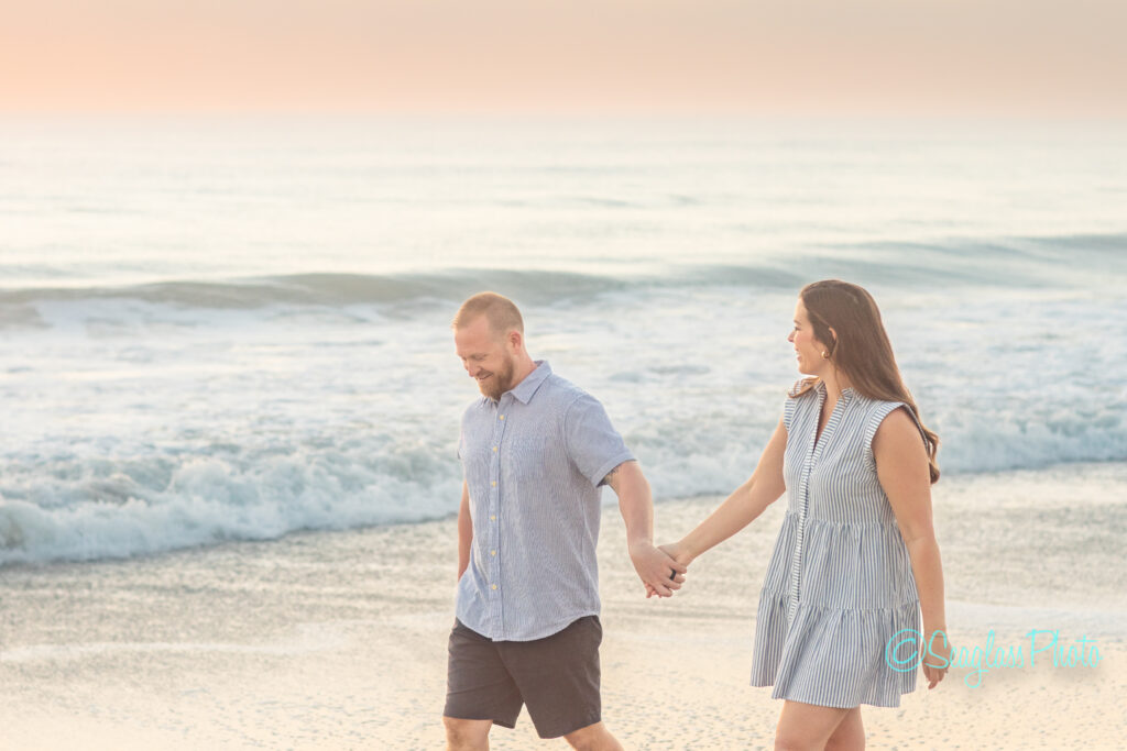 A couple walks hand in hand along the beach at sunset. The man wears a blue shirt and dark shorts, while the woman wears a striped dress. Waves gently roll onto the shore in the background under a pastel-colored sky. Vero Beach Photoshoot