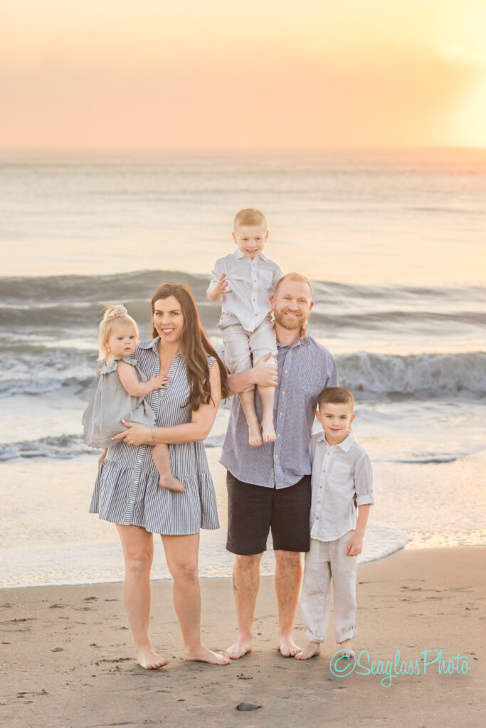 A family of five stands on the beach at sunset. The woman holds a young girl in a gray dress. Two boys and a man, wearing light-colored clothing, smile at the camera. Waves gently lap at the shore behind them. Vero Beach Photoshoot