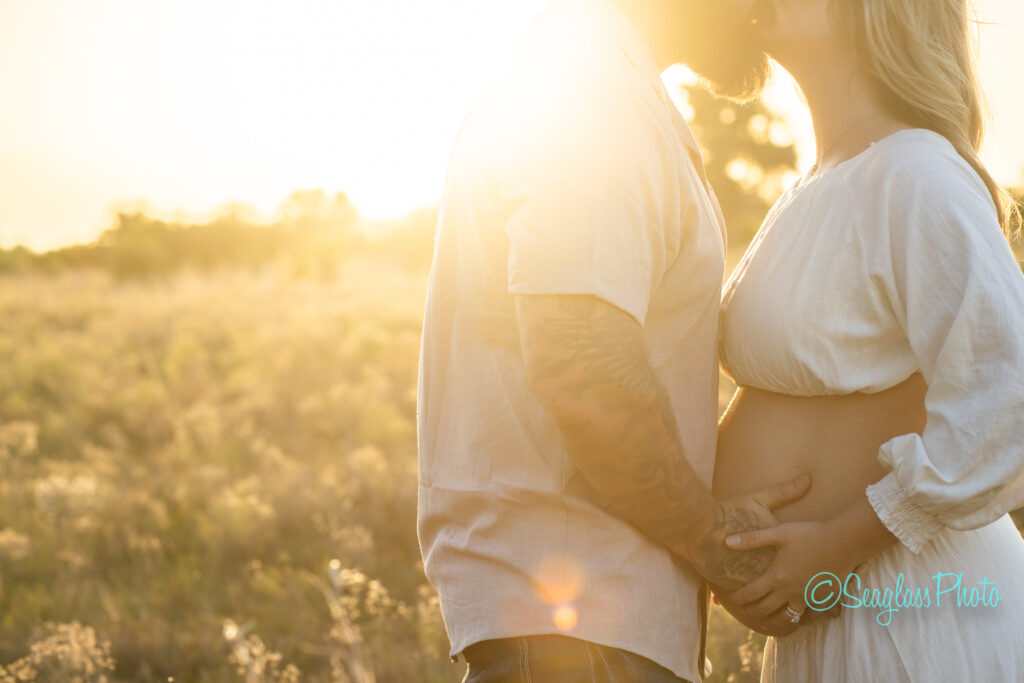 A couple embracing in a sunlit field. The womans hand is on her pregnant belly, while the mans hand rests nearby. They appear to be sharing an intimate moment amidst a golden glow.