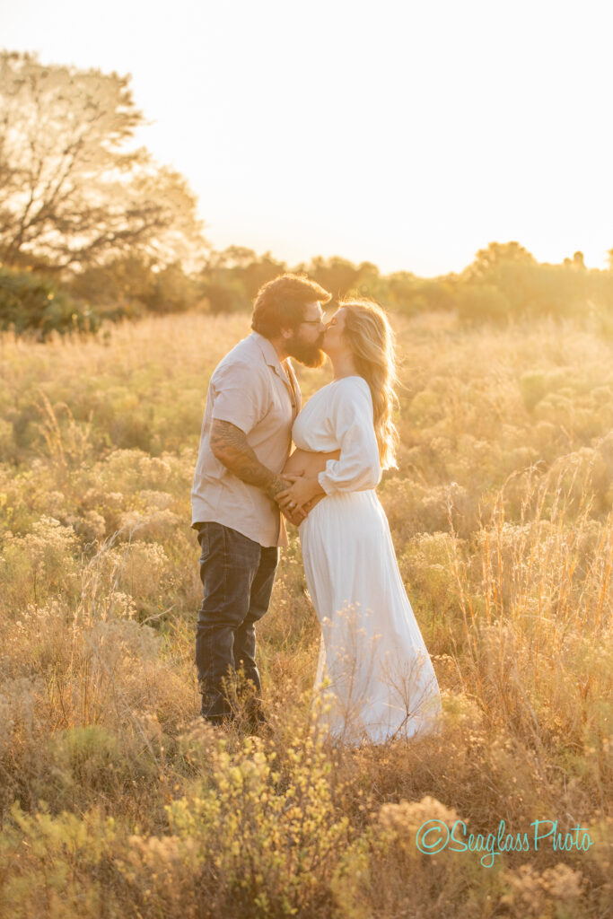 A couple stands in a sunlit field, both wearing light-colored clothing. The man, with a beard, gently kisses the woman on the forehead. She is wearing a white dress and holding her belly, indicating she is pregnant. The scene is warm and serene.
