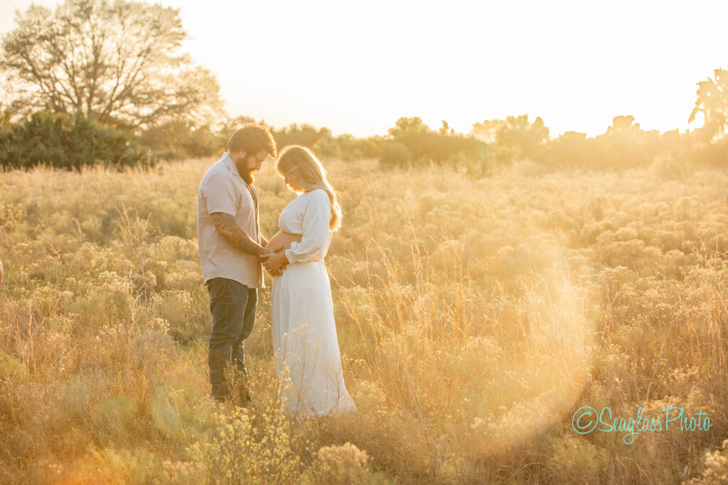 A couple stands closely in a sunlit field, surrounded by tall grass. The woman wears a long white dress, and the man is in a light shirt and pants. They appear to be holding hands. A large tree is visible in the background, and the sun creates a warm glow.