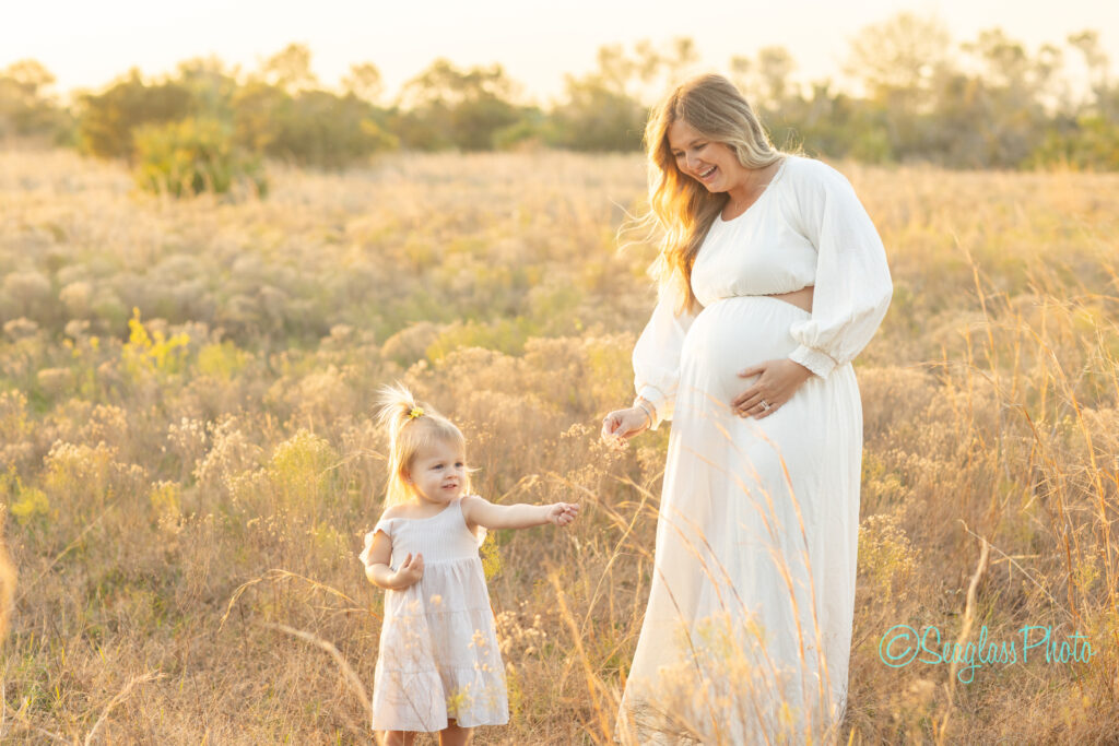 A pregnant woman in a white dress stands in a field, smiling at a young girl beside her. The girl, in a white dress, reaches out toward flowers. The sun sets in the background, casting a warm glow over the scene.