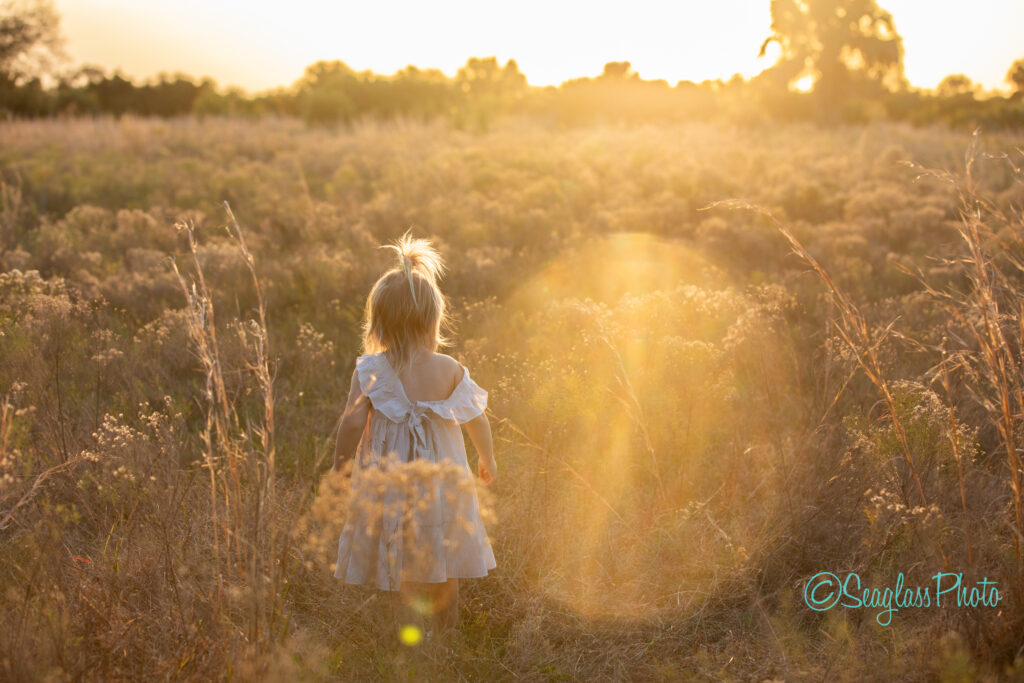 A young child with light hair stands in a sunlit field of tall grass and wildflowers, wearing a white dress. The golden light of the setting sun creates a warm, dreamy atmosphere with a lens flare effect.