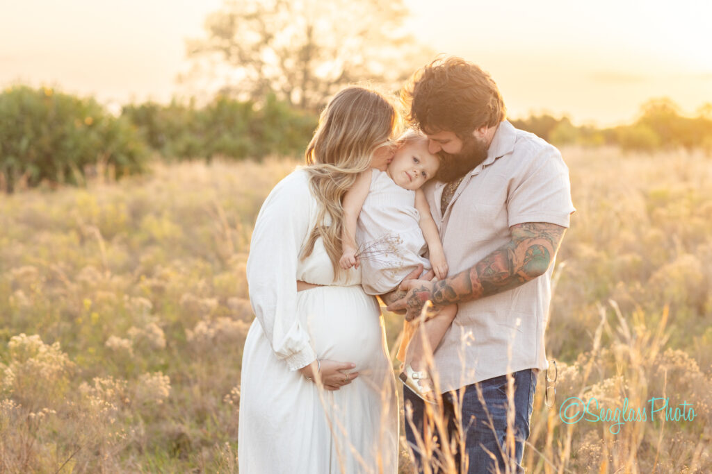 A pregnant woman in a white dress stands in a field at sunset, cradling her belly. A man with tattoos holds a child, who rests their head on her shoulder. They are embracing, with warm light surrounding them.