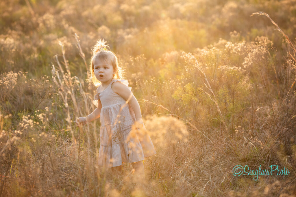 A young child with light hair stands in a sunlit field, surrounded by tall grasses and wildflowers. The child wears a sleeveless dress and looks towards the camera, with the sun creating a warm glow around them.