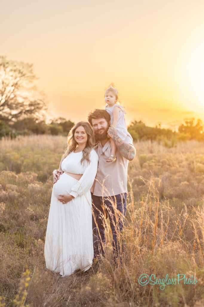 A pregnant woman in a white dress stands in a field with a bearded man holding a child on his shoulders. The sun sets behind them, casting a warm glow over the scene.