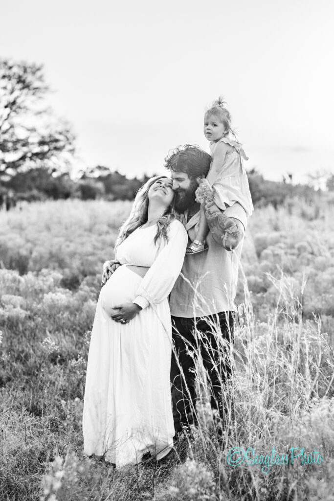 A black and white photo of a smiling pregnant woman standing in a field, holding her belly. A man beside her, with a child on his shoulders, affectionately looks at her. The field has tall grass and trees in the background.
