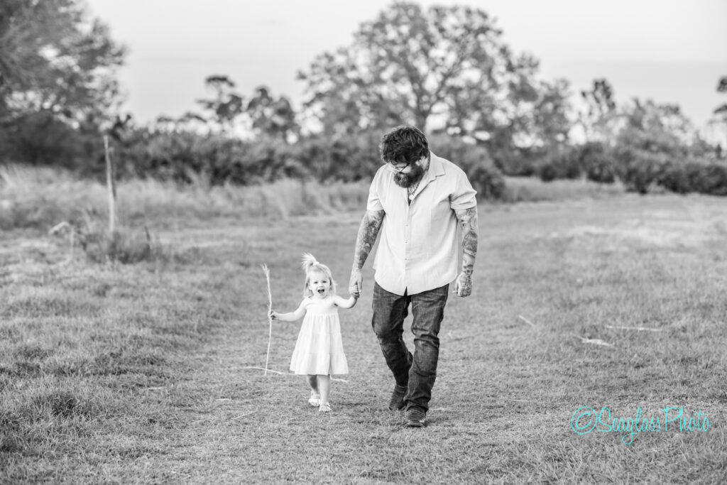 A bearded man with tattoos walks hand in hand with a young girl in a white dress, who is joyfully holding a stick. They are on a grassy path surrounded by trees. The image is in black and white.