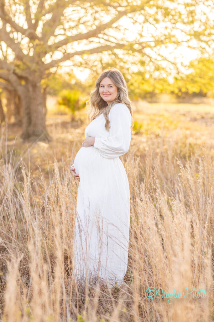 A pregnant woman in a white dress stands in a sunlit field, surrounded by tall grasses. She gently holds her belly and smiles softly, with a large tree in the background and golden light filtering through the branches.