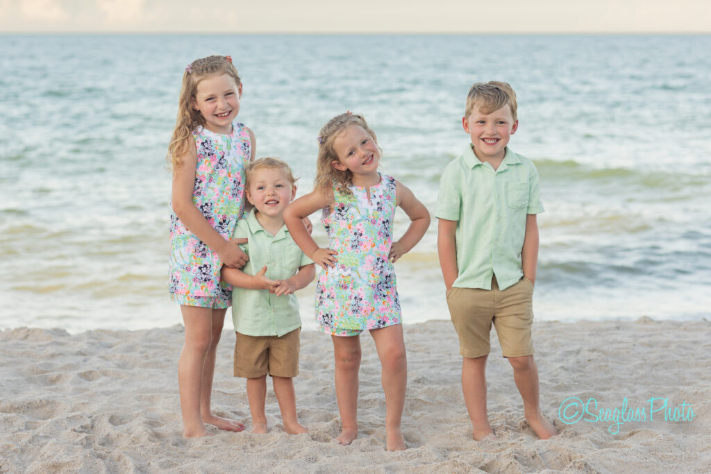 Four young children stand smiling on a sandy beach with the ocean in the background. The two girls wear matching floral dresses, while the two boys wear mint green shirts and tan shorts. Vero Beach Photoshoot