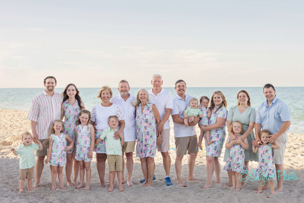 A large family poses together on a sandy beach, all wearing coordinated pastel and floral outfits. The ocean and sky are visible in the background, and several children stand or are held by adults, smiling at the camera. Vero Beach Photoshoot