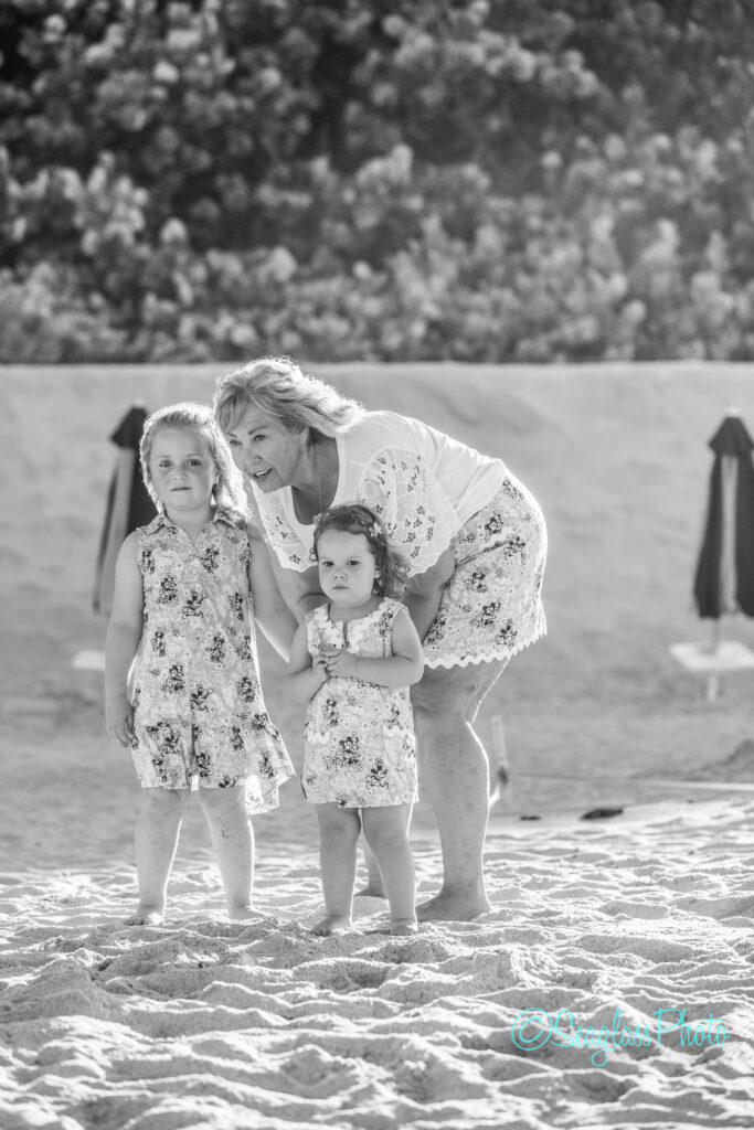 A woman kneels in the sand beside two young girls, all wearing matching floral outfits. The girls stand close together, looking ahead, with trees and sun umbrellas blurred in the background. The image is in black and white. Vero Beach Photoshoot