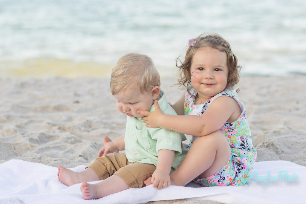 A young girl sitting on a beach towel gently pinches a baby boy’s cheek while smiling at the camera. The boy looks down, and the ocean and sand are visible in the background. Vero Beach Photoshoot