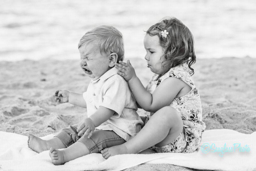 A young girl sitting on a blanket at the beach gently holds a crying toddler boy’s face, trying to comfort him. The photo is in black and white. Vero Beach Photoshoot