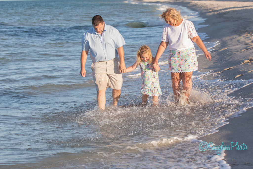 A man, woman, and young girl hold hands while walking through shallow waves along a sandy beach, smiling and enjoying the sunny day together. Vero Beach Photoshoot