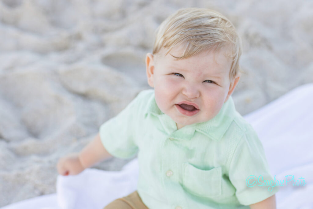 A young blonde toddler in a light green shirt sits on a white blanket at the beach, looking up and smiling with sand visible in the background. Vero Beach Photoshoot