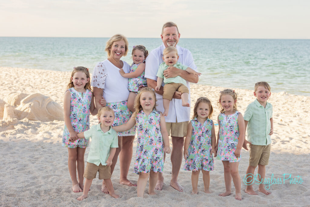 A smiling couple stands on a sandy beach with eight young children, all dressed in coordinated pastel outfits. The ocean and sky are in the background, and a sand sculpture is visible on the left. Vero Beach Photoshoot