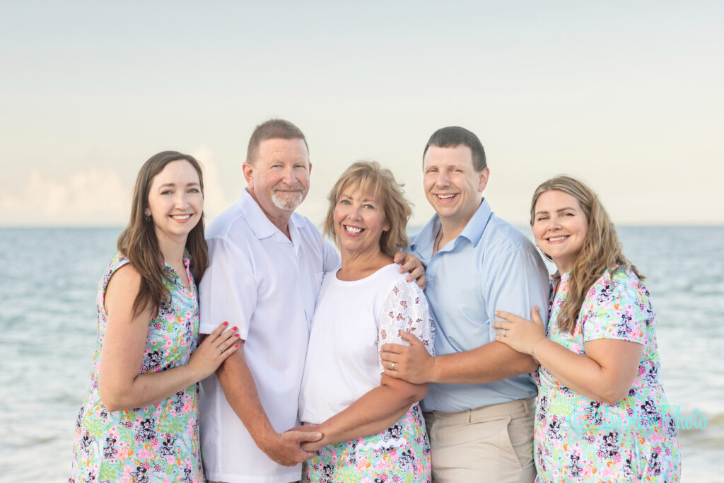 A family of five poses and smiles together on a beach with the ocean in the background. The women wear floral dresses, and the men wear light-colored shirts. The weather is clear and sunny. Vero Beach Photoshoot