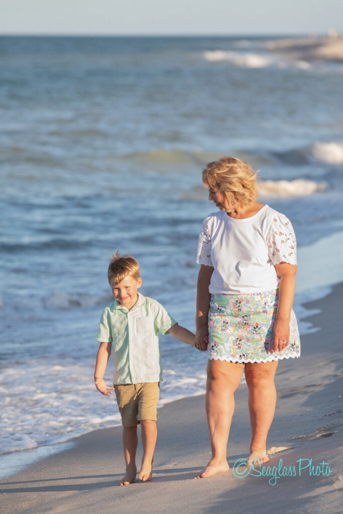 An older woman and a young boy walk hand in hand along the shoreline on a sunny day. They both smile and look relaxed. Gentle waves roll onto the sand beside them. Vero Beach Photoshoot