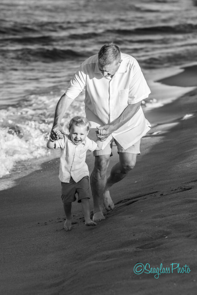 A smiling older man holds a young boy’s hands as they walk along the shoreline, waves in the background. Both are barefoot and dressed in light clothing, enjoying a sunny day at the beach. Vero Beach Photoshoot