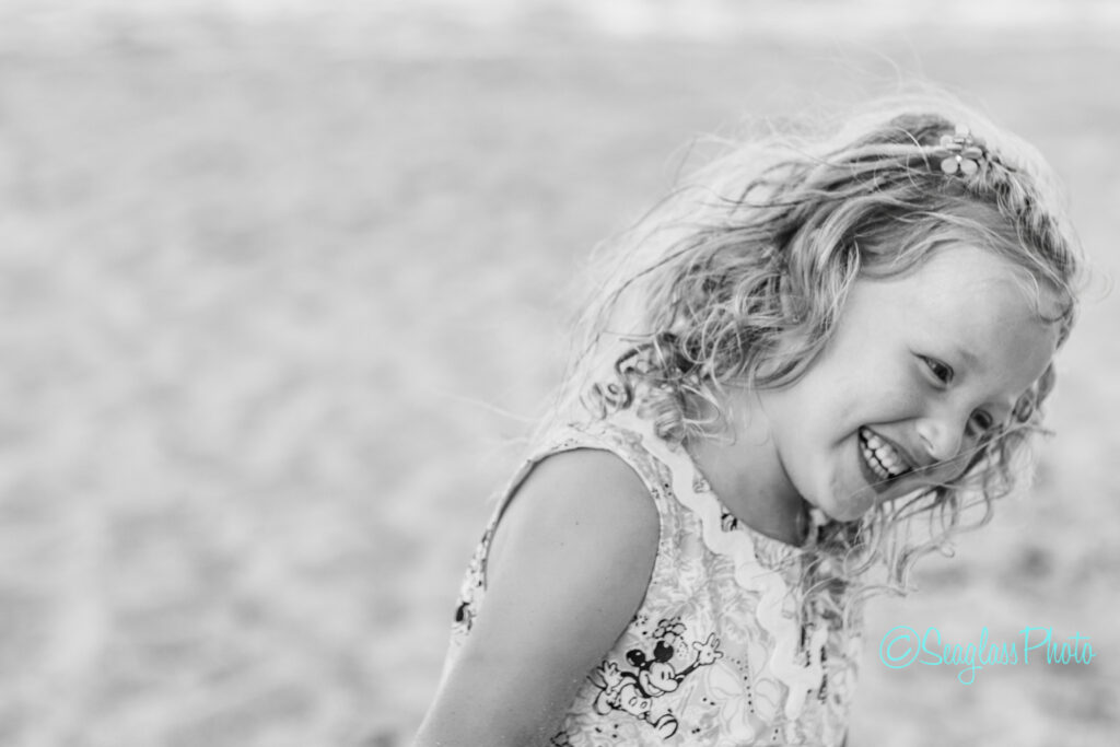 A young girl with curly hair, wearing a patterned dress, smiles and laughs while looking to the side. The background is blurry, suggesting she is outdoors, possibly on a beach. The photo is in black and white. Vero Beach Photoshoot