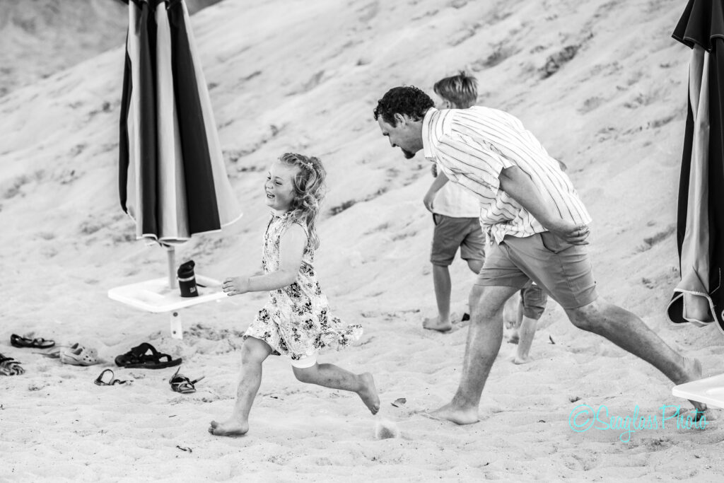 A man and two children play and run on a sandy beach near large umbrellas. The little girl in a floral dress smiles as the man chases her. Shoes are scattered on the sand. The scene is bright and joyful. Vero Beach Photoshoot