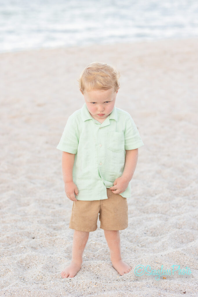A young boy with blonde hair, wearing a light green shirt and tan shorts, stands barefoot on a sandy beach, looking down with a serious expression. The ocean is visible in the background. Vero Beach Photoshoot