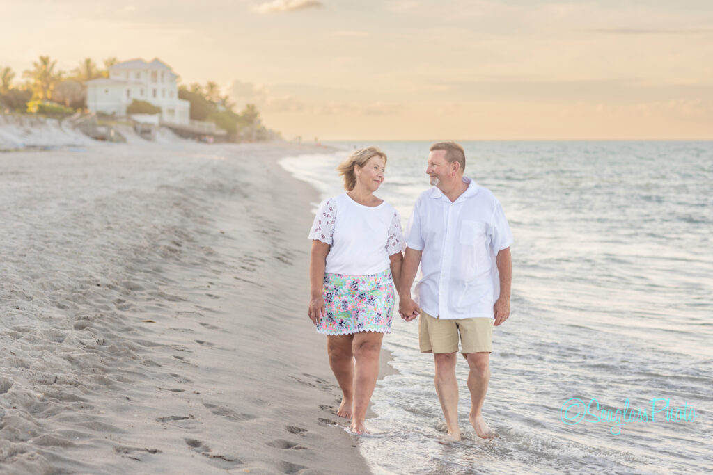 A middle-aged couple walks hand-in-hand along a sandy beach at sunset, smiling at each other, with gentle waves and houses visible in the background. Vero Beach Photoshoot