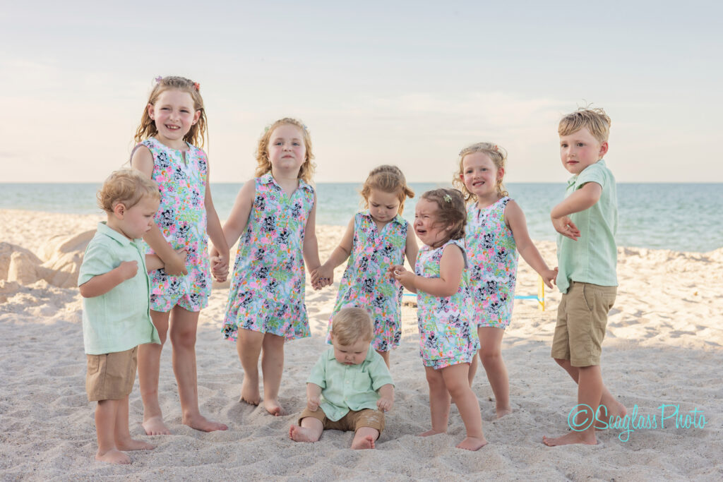 Eight young children stand and play on a sandy beach. Most of the girls wear matching floral dresses, while the boys wear light green shirts and khaki shorts. The ocean and blue sky are visible in the background. Vero Beach Photoshoot