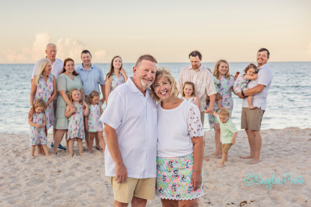 A large multigenerational family poses on a sandy beach at sunset, smiling at the camera. An older couple stands in front, with children and adults in colorful outfits gathered behind them near the ocean. Vero Beach Photoshoot