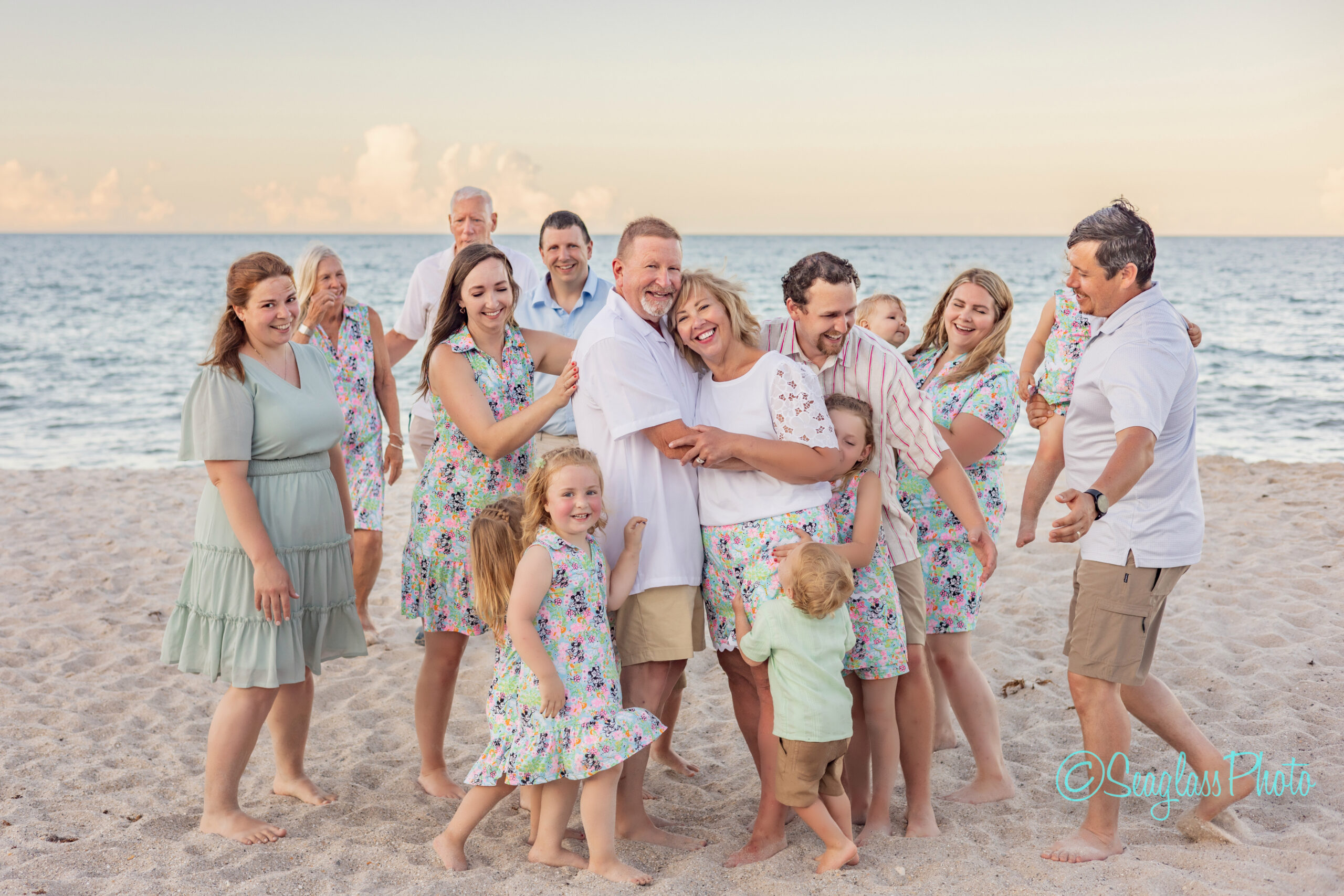 A large multigenerational family smiles and laughs together on a sandy beach at sunset, with adults and children dressed in coordinating pastel outfits, the ocean and sky in the background. Vero Beach Photoshoot