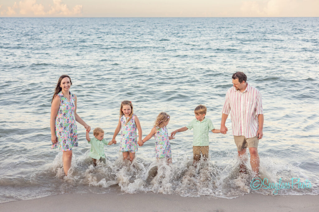 A family of five, including two adults and three young children, holds hands and stands at the shoreline as gentle waves splash around their feet. They are dressed in light, summery clothes and smile at the camera. Vero Beach Photoshoot