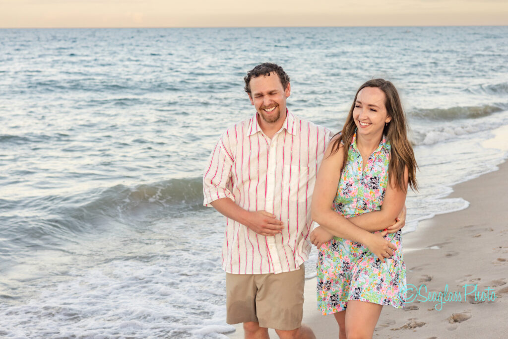 A smiling man and woman walk together along the shoreline at a beach, with gentle waves and a calm sea in the background. The man wears a striped shirt and shorts; the woman wears a floral dress. Vero Beach Photoshoot
