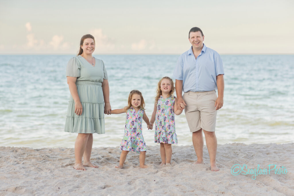 A family of four stands barefoot on a sandy beach, smiling at the camera. The parents hold hands with their two young daughters. The ocean and sky are visible in the background. Vero Beach Photoshoot