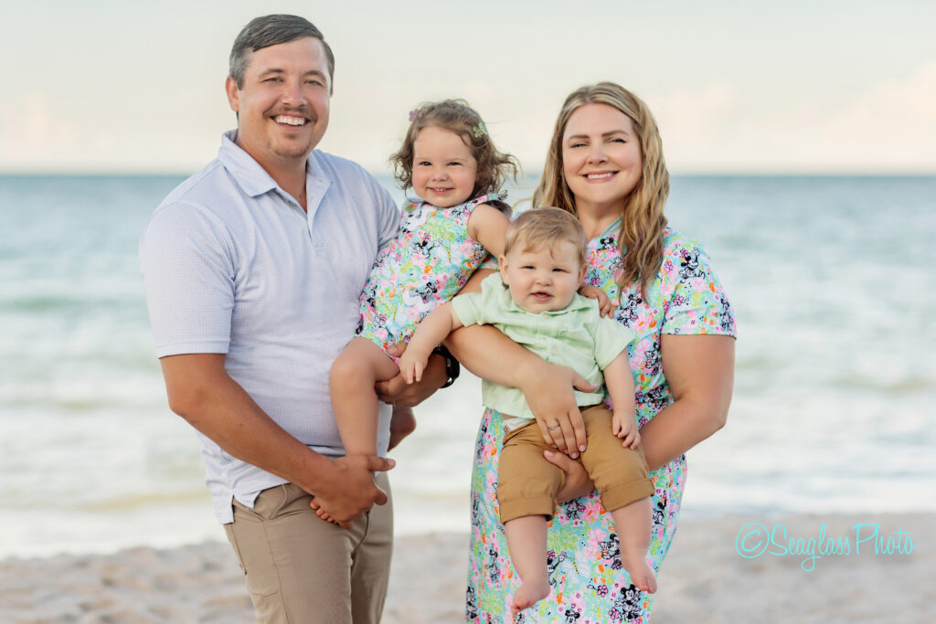 A smiling family of four stands on a sandy beach with the ocean in the background. The parents hold a young girl and toddler boy, all dressed in light, summery clothes. The weather is clear and bright. Vero Beach Photoshoot