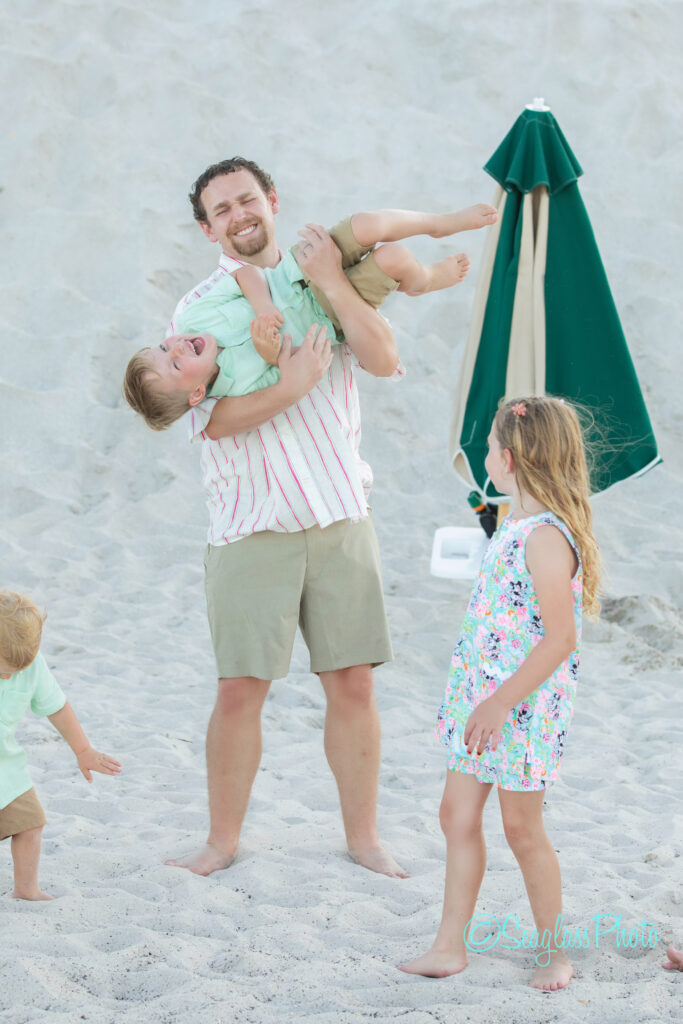 A man on a sandy beach smiles while holding a laughing boy sideways. A girl in a floral dress and another small child play nearby. An upright green beach umbrella is in the background. Vero Beach Photoshoot