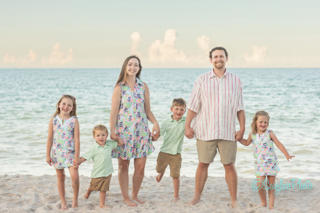 A smiling family of five, including two girls, two boys, and their parents, stands barefoot on a sandy beach in front of the ocean, wearing pastel summer outfits. Vero Beach Photoshoot