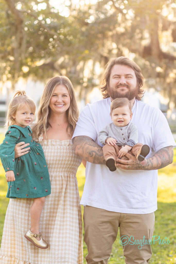 A smiling family of four stands outdoors in sunlight. The mother holds a young girl in a green dress. The father, with tattoos and a beard, holds a baby boy in grey and brown. Trees and sunlight brighten the background. Vero Beach Photoshoot