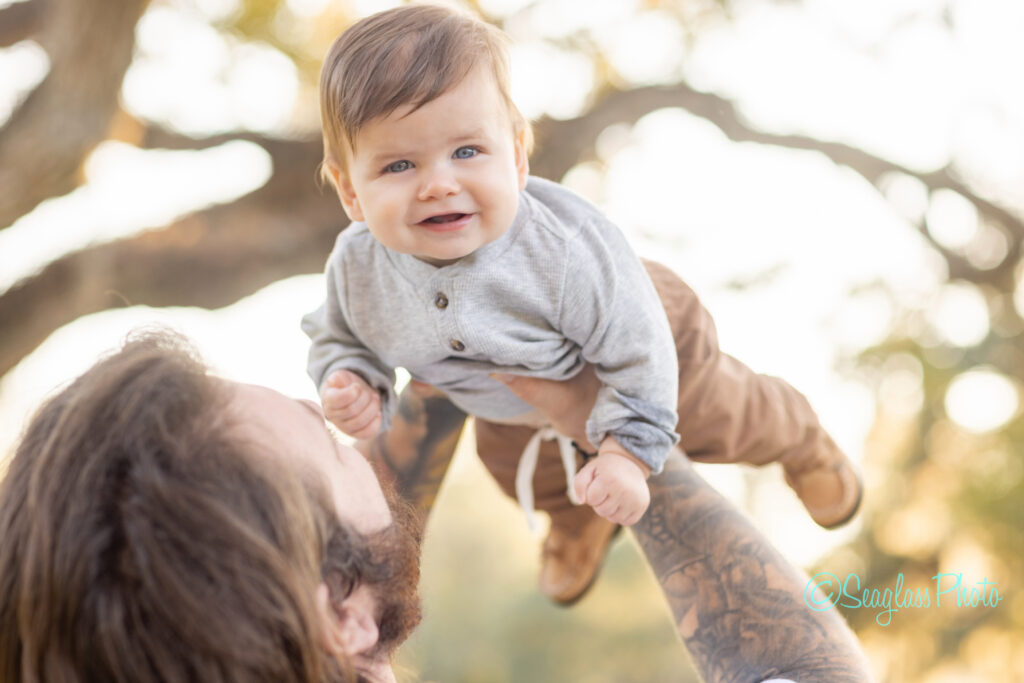 A bearded man with tattoos lifts a smiling baby boy in brown pants and a gray shirt into the air outdoors, with sunlight filtering through blurred trees in the background. Vero Beach Photoshoot