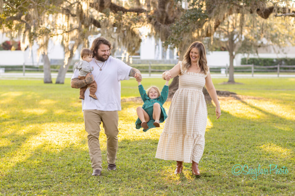 A smiling family walks in a sunlit park. The father holds a toddler and swings a laughing child, while the mother, in a gingham dress, holds the other hand. Trees with hanging moss are in the background. Vero Beach Photoshoot