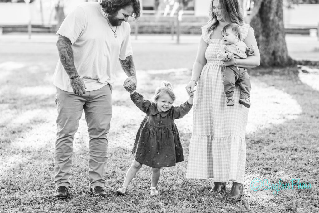 A man and woman hold hands with a smiling young girl between them, while the woman also holds a baby. They stand together outdoors on grass under a tree, all appearing happy. The photo is in black and white. Vero Beach Photoshoot