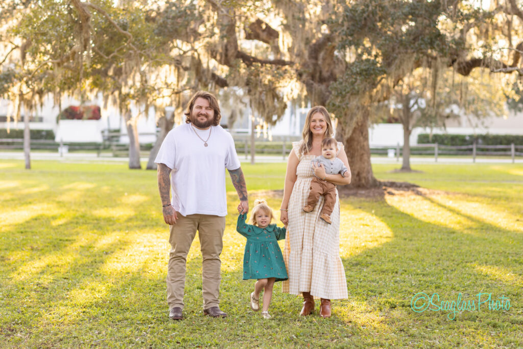 A family of four stands on grass under trees. The father and young daughter hold hands; the mother holds a toddler. Sunlight filters through the trees, casting long shadows. Everyone is smiling and casually dressed. Vero Beach Photoshoot
