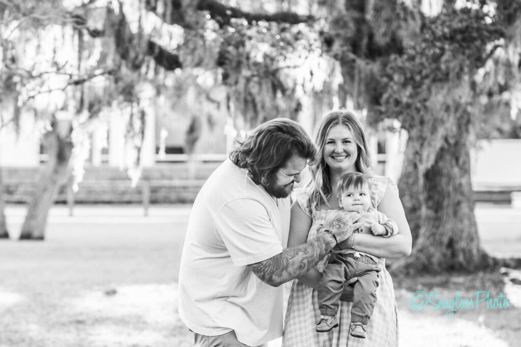 A man and woman stand outdoors, smiling, while holding a baby. The man looks at the baby and the woman faces the camera. Trees with hanging moss are in the background. The photo is black and white. Vero Beach Photoshoot