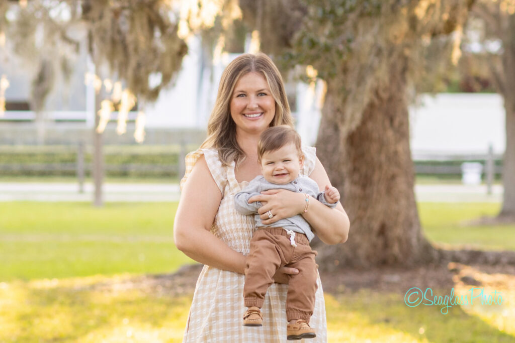 A smiling woman in a light plaid dress stands outdoors holding a happy baby boy dressed in a gray shirt and brown pants. They are in a sunny park with green grass and large trees in the background. Vero Beach Photoshoot
