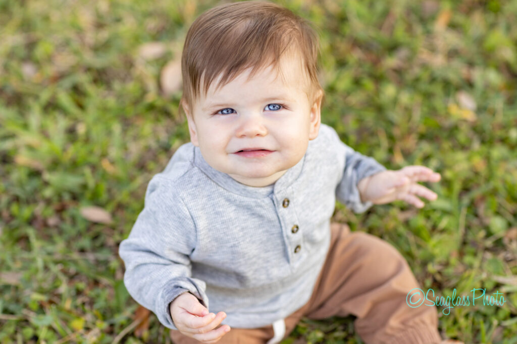 A baby with light brown hair and blue eyes sits on grass, wearing a gray long-sleeve shirt and brown pants, looking up and smiling slightly at the camera. Vero Beach Photoshoot
