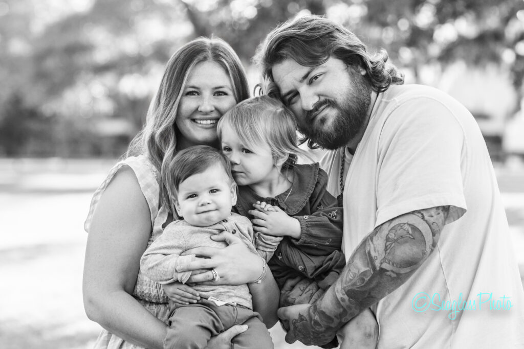 A smiling family of four poses outdoors. The mother holds a baby while the father gently hugs an older child, who is nuzzling close. Trees and sunlight create a soft, natural background. Vero Beach Photoshoot