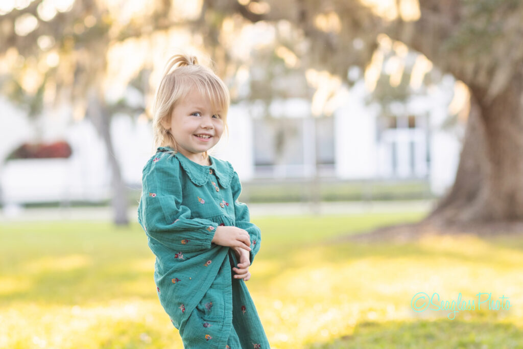 A young girl with blonde hair in a ponytail, wearing a teal dress with floral patterns, smiles and holds her dress while standing on grass in a sunlit park with blurred trees in the background. Vero Beach Photoshoot