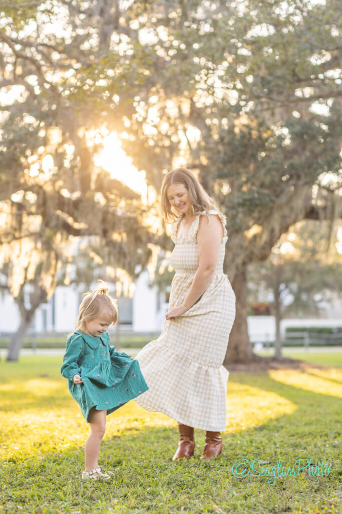 A woman in a light plaid dress and boots smiles while holding her dress, watching a young girl in a teal dress playfully lift her foot outdoors, with sunlight streaming through trees in the background. Vero Beach Photoshoot